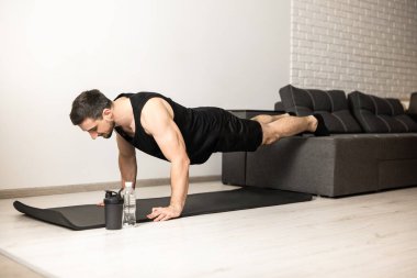 Sportif adam kanepede bacaklarıyla zeminden itiyor. Siyah yoga minderiyle egzersiz. Siyah giysili adam evde egzersiz yapıyor. Arka planda beyaz modern oda. Formda olmak için idman yapan güçlü bir adam..