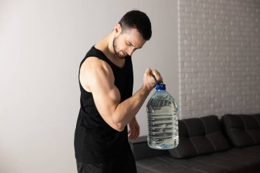 Güçlü adam evinde büyük bir şişe suyla buzağı yetiştirme egzersizleri yapıyor. Geniş ve parlak dairesinde, minimalist bir iç mekanı var. Siyah spor kıyafetli bir adam sabah antrenmanı yapıyor..