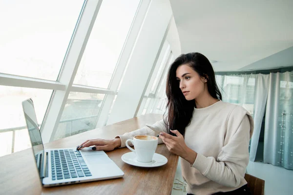 Freelancer woman working on laptop and typing on smartphone to customer. White room on background. Woman programmer looking for a bug in code and searches for examples on web using a phone.