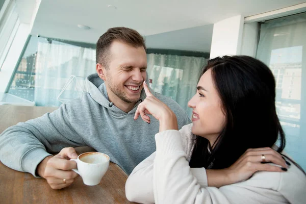 Χαρούμενο ζευγάρι που περνάει καλά με ένα φλιτζάνι καφέ. Ευτυχισμένη μελαχρινή γυναίκα να δείχνει από το δάχτυλό της τη μύτη του άντρα και να χαμογελάει. Άνθρωπος που αλληθωρίζει και κρατάει ένα φλιτζάνι καπουτσίνο. — Φωτογραφία Αρχείου