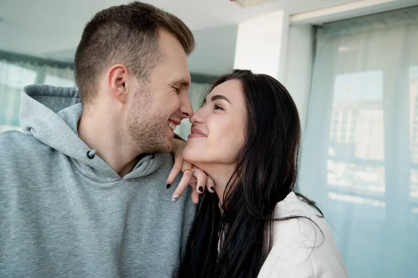 Beautiful couple in love embraced and gently rubs noses. Brunette woman with long hair looking deeply into mans eyes. White light room on background. Young cheerful couple touched one anothers nose. — Stock Photo, Image