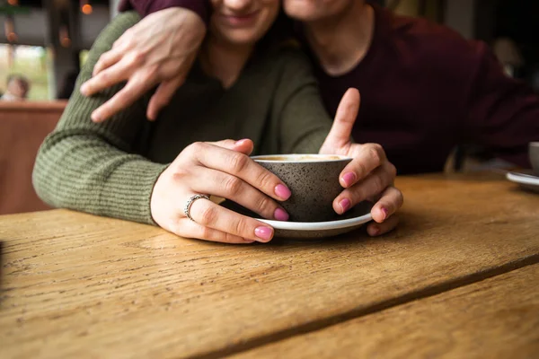 Blurred young couple in cafe. Woman holding cup of warm coffee when man embraces her. Cozy atmosphere in coffee shop. Cup of cappuccino or flat white. Happy couple hugging in coffee shop. — Stock Photo, Image