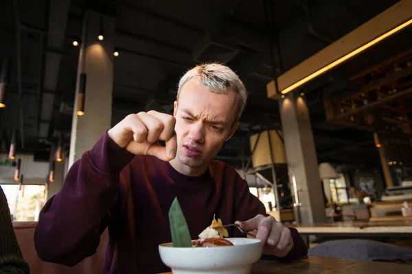 Disgusted young man found hair in his meal at cafe and looks at it with loathing. Bad customer service concept. Blond young man in casual clothes isnt satisfied with the food.