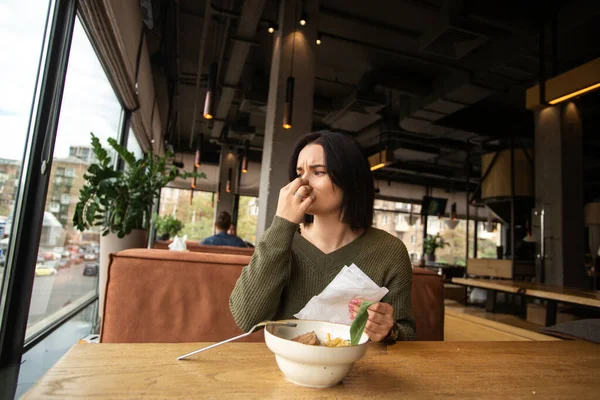 Young woman covers her nose because of bad smelling food. Terrible meal concept. Brunette woman understand that she doesnt feel smells. Coronavirus concept. Public place on background.