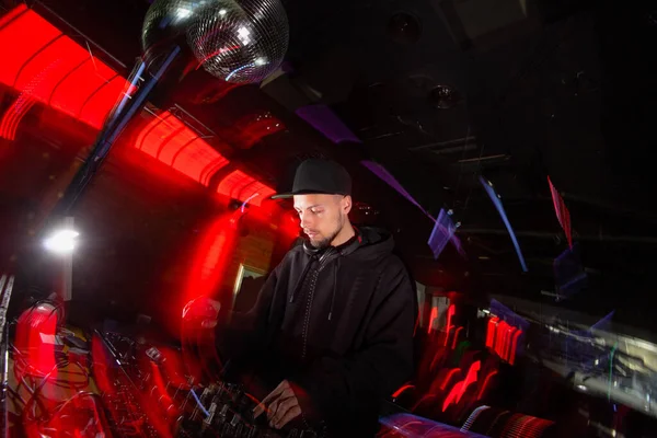 Concentrated DJ plays music on a party. Good looking young man in black hat and casual black clothes uses turntable to mix music. Blurred background with red light.