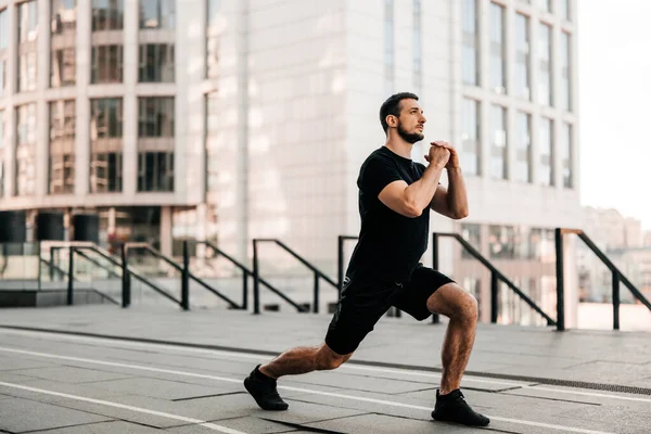 Formda adam ileri hamle yapıyor. Büyük şehir konseptinde spor. Güçlü adam koşmadan önce bacaklarını esnetiyor. Sağlık sigortası konsepti. Siyah spor giyimli bir koşucu. Kentsel spor konsepti.