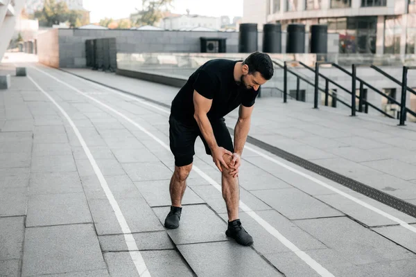 Attractive man feeling pain in his foot during sport workout outdoors. Male runner in black sportswear and sneakers standing and feeling cramps in his left leg. Urban sport concept.