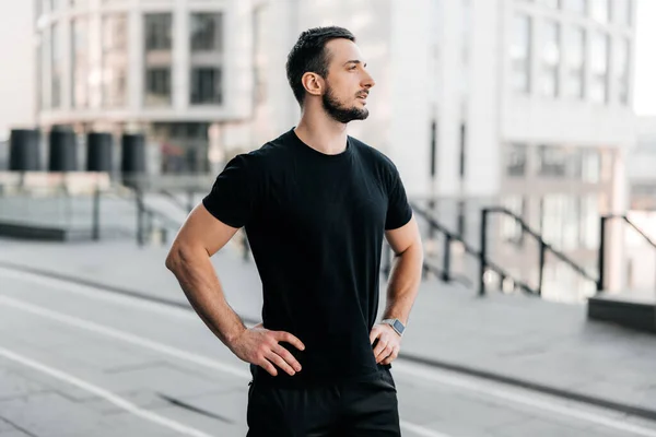 Erkek koşucu şehrin güzel sabah manzarasına hayran olmak için koşuya ara verdi. Siyah spor kıyafetli bir adam ellerini belinden tutup başka tarafa bakıyor. Şehir sporu konsepti. Sabah sporu.