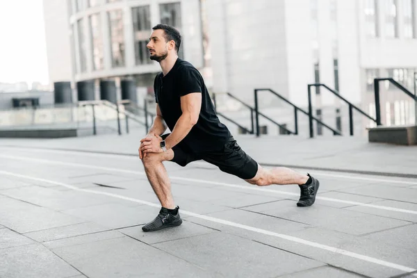 Un hombre fuerte se lanza hacia adelante. Vista lateral. Deporte en concepto de gran ciudad. Ajuste hombre estirando las piernas antes de trotar. Corredor en ropa deportiva negra calentándose antes del entrenamiento. Concepto de deporte urbano. — Foto de Stock