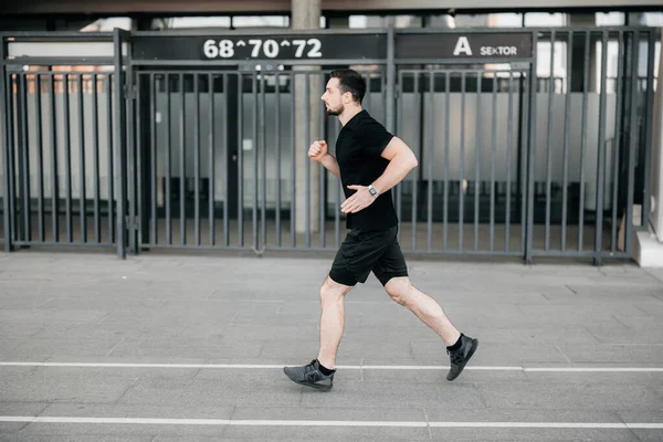 午前中に集中的に男性のランナーの抜粋。都市生活の概念。黒のスポーツ服やスニーカーのアスレチックマンは屋外でジョギング。健康的なライフスタイル。走ってる男。活発な生活. — ストック写真