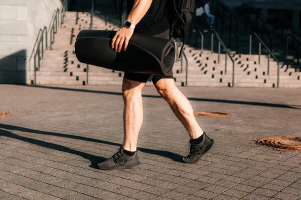 Tired and satisfied male sportsman returns home after morning workout. Cut view of sporty mans legs. Urban sport concept. Man walking on street and holding rolled yoga mat.