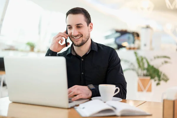 Úspěšný pracovník na dálku. Mladý svobodný muž, spokojený s telefonátem. Kavárna na pozadí. Muž na volné noze, programátor pracující v kavárně. Rád si promluví s klientem.. — Stock fotografie