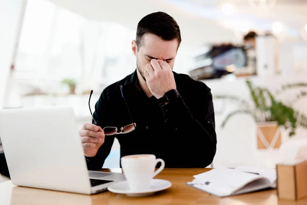 Dê um descanso aos seus olhos Jovem freelancer cansado de camisa preta sofre de dor e secura nos olhos. Ele trabalha muito ambiente aconchegante cafetaria no fundo. Copo de cappuccino gostoso na mesa. — Fotografia de Stock