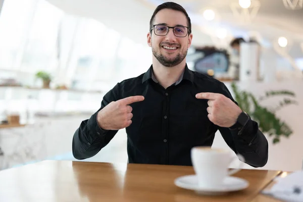 Ahoj, to jsem já Atraktivní veselý muž v černé košili, ukazující si prsty na hrudi a usmívající se. Sedí v kavárně a čeká na setkání s online přáteli. — Stock fotografie