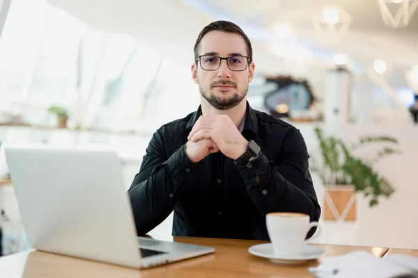 Sebevědomý atraktivní muž hledící do kamery. Moderní notebook a šálek cappuccino na dřevěném stole. Lehký elegantní kavárna interiér na pozadí. Muž na volné noze pracující v kavárně. — Stock fotografie