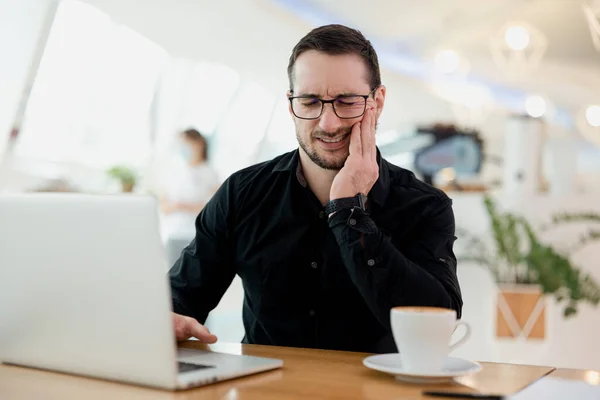 Conceito de dor de dente. Homem jovem freelancer sofrendo de dor de dente, cárie dentária ou sensibilidade. Visite seu dentista regularmente Homem de camisa preta cobrindo sua bochecha. laptop moderno e café na mesa. — Fotografia de Stock