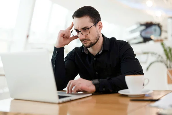 Inteligente jovem profissional pensando sobre a tarefa técnica. Um homem concentrado a segurar a cabeça e a usar o portátil. Conceito de trabalho remoto. Xícara de cappuccino gostoso. — Fotografia de Stock