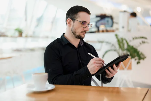 Mužský webdesigner nebo umělec kreslí na svém tabletu. Digitální umělecký koncept. Mladý soustředěný muž s vousy a brýlemi pomocí svého přístroje v kavárně. — Stock fotografie