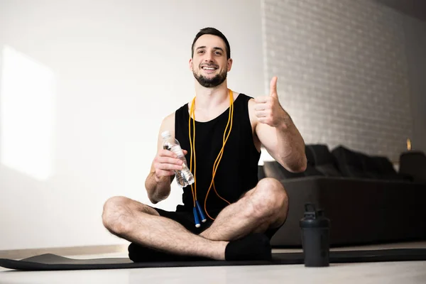 Un joven haciendo ejercicio en casa. Guy se sienta solo en el mapa de yoga negro, sostiene la botella de agua y los pulgares hacia arriba. Amarillo saltar la cuerda alrededor del cuello. Descanso atlético y relax. Pequeño descanso. — Foto de Stock