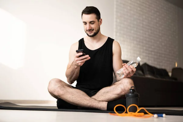 Hombre descansando después del entrenamiento en casa con agua y teléfono. Joven hombre deportivo después de practicar yoga, romper en hacer ejercicio, relajarse en la estera de yoga, mensajes de texto en el teléfono inteligente. Concepto sanitario. — Foto de Stock