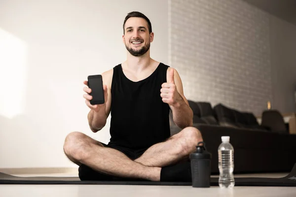 Hombre satisfecho pulgares hacia arriba y muestra los deportes favoritos aplicación móvil. Ejercicio concepto cotidiano. Dos botellas deportivas de agua. El hombre feliz se sienta en una esterilla de yoga negra con piernas cruzadas. Concepto de estilo inteligente. — Foto de Stock