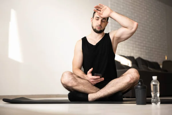 Hombre musculoso limpia el sudor de su frente después de un duro entrenamiento en casa. Concepto de estilo de vida saludable. Hombre cansado después de un entrenamiento intensivo. Botella de agua de plástico. Sala de estar luminosa sobre fondo. — Foto de Stock