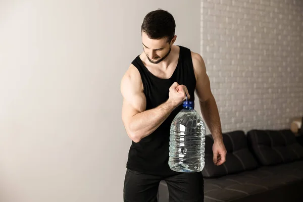 Hombre atlético haciendo ejercicios de aumento de pantorrilla con una gran botella de agua. Habitación blanca moderna en el fondo. Hombre fuerte en ropa deportiva negra haciendo ejercicio para tener un cuerpo en forma. — Foto de Stock