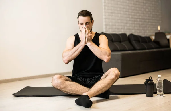 Un hombre enfermo con secreción nasal sentado en una esterilla de yoga negra. Quédate en casa concepto. El joven moreno se sonó la nariz después del entrenamiento. Practicar deportes en casa si se siente enfermo. — Foto de Stock