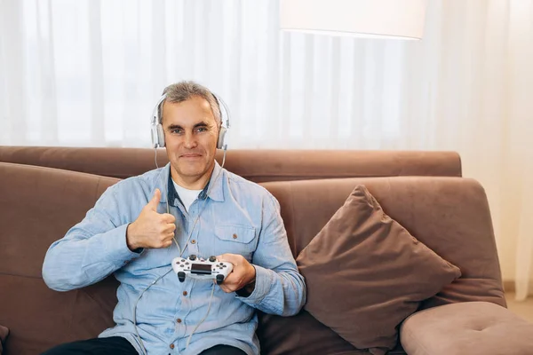 Mature gamer man playing video game using joystick and headphones happy with big smile, thumbs up. Excellent sign. Living room on background. Man in casual clothes.