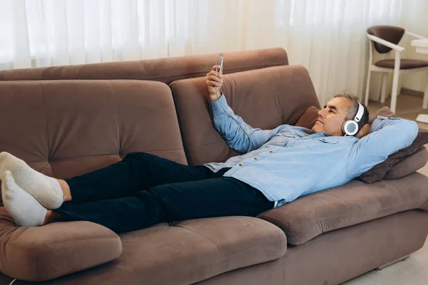 Descanse en casa después del trabajo durante la cuarentena. Hombre maduro con el pelo gris se encuentra en el sofá y escucha música en su teléfono inteligente. Tiempo libre. Concepto de tecnología moderna. — Foto de Stock