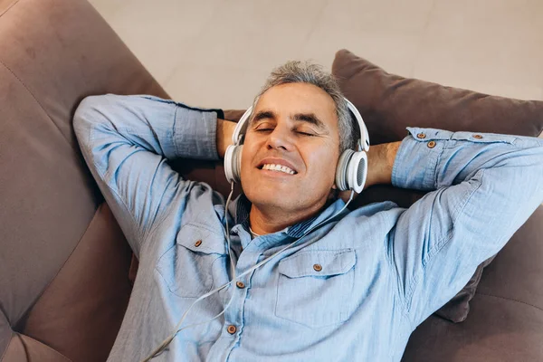 Uomo di mezza età che ascolta musica, sdraiato sul divano con gli occhi chiusi e sorridente con i denti. Stile casual. Uomo godendo la sua musica preferita e rilassante a casa. Trascorrere del tempo durante la quarantena. — Foto Stock