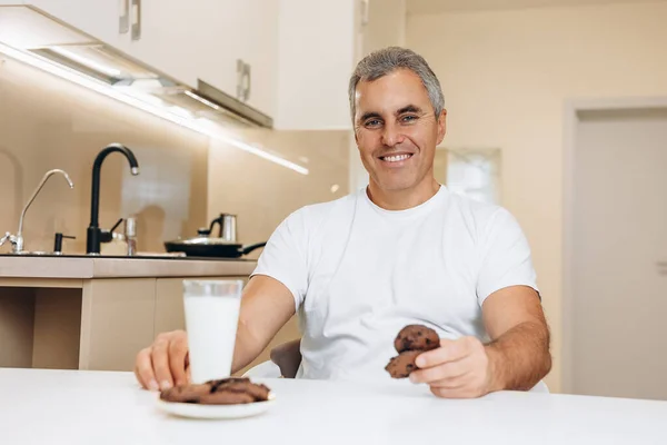 Breakfast concept. Cheerful senior man in white t-shirt has a glass of milk and chocolate cookies for a breakfast today. Smart kitchen interior with light furniture.