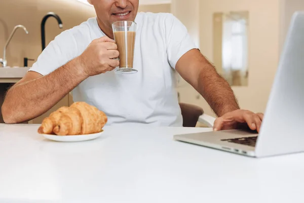 Satisfied man drinks latte or coffee with milk and uses his modern laptop. Crispy croissant on a white table. Man watching funny videos and enjoying breakfast. Modern kitchen on background.