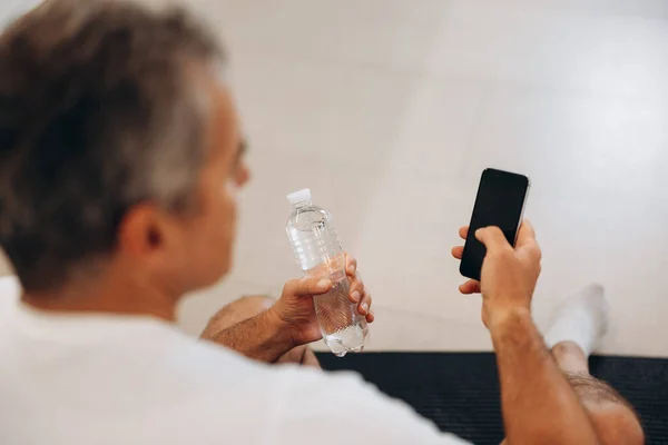 Hombre mayor usando su teléfono inteligente y sosteniendo la botella de agua de plástico. Por encima del hombro. Hombre en camiseta blanca sentado en estera de yoga negro y descansando después de una dura práctica. — Foto de Stock