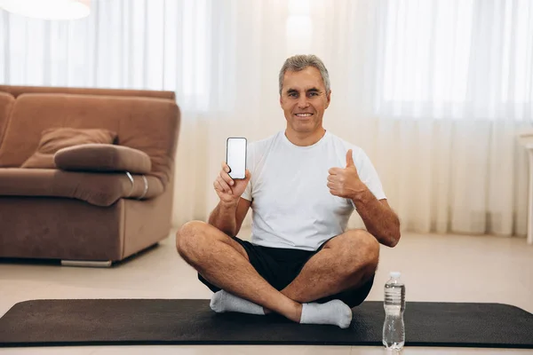 Літня людина дійсно любить свій улюблений додаток для тренування. Старий тримає свій смартфон, піднімаючись і сидячи з перехресними ногами. Дивовижний телефонний додаток. Чоловік задоволений його практикою йоги . — стокове фото