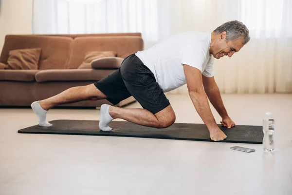 Elderly man doing mountain climber exercises on black yoga mat. Morning workout. Modern living room on background. Plastic bottle of water. Hard workout. Training at home. Elderly sport concept.