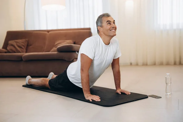 Yaşlı adam oturma odasında kobra pozu verirken meditasyon yapıyor. Esneme egzersizleri. Kapalı alanda yoga yapan mutlu bir adamın fotoğrafı. Sabah sporu. Plastik su şişesi. Güçlü kollar.