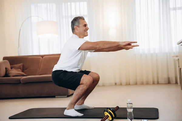 Antrenman zamanı. Güçlü kıdemli adam evinde hafif oturma odasında çömelme egzersizi yapıyor. Sağlıklı yaşam tarzı, refah ve aktivite konsepti. Siyah ve beyaz spor giyim. Aktif ve mutlu yaşlılık..