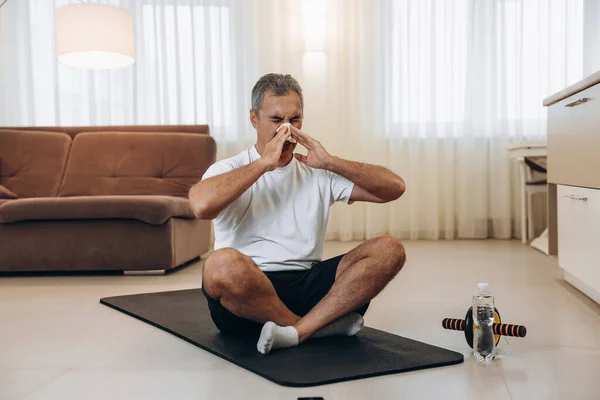 Burnu akan yaşlı hasta adam siyah yoga minderinde oturuyor. Evde kal konsepti. Adam spor yaptıktan sonra burnunu siliyor. Miden bulanırsa evde spor yapıyorsun. Spor malzemeleriyle egzersiz.