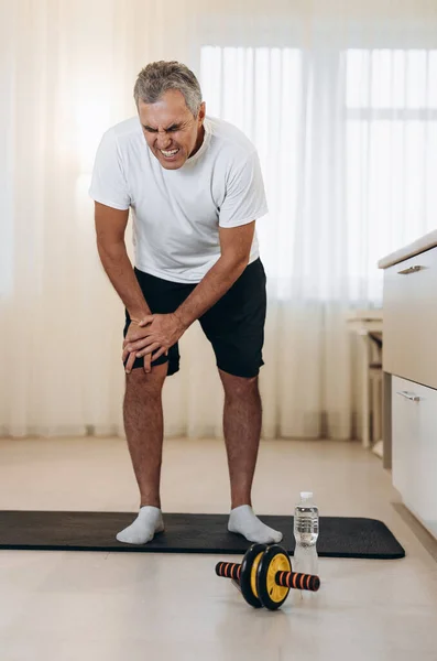 Evde spor yaparken bacağında ağrı hisseden yaşlı bir adam. Siyah beyaz spor kıyafetli erkek sporcu yoga minderinin üzerinde duruyor ve sağ bacağına kramplar giriyor..