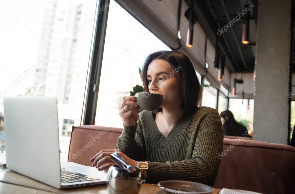 Freelancer working in cozy atmosphere concept. Concentrated young woman drinks coffee from grey cup and looks into laptop on her table with attention. Big well illuminated windows on background.