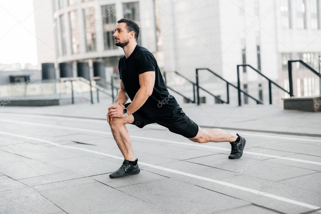 Strong man doing lunge forward. Side view. Sport in big city concept. Fit man stretching legs before jogging. Runner in black sportswear warming up before workout. Urban sport concept.