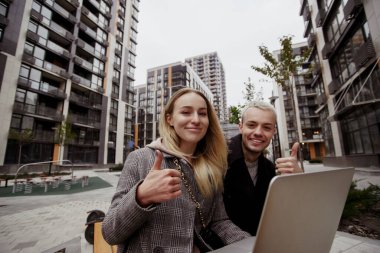 Genç adam ve kadın dışarıda çalışmayı seviyorlar. Çift bankta oturuyor ve başparmak kaldırıyor. Arka planda modern apartman blokları. Çekici bir kadın ve yakışıklı bir adam gülümseyip kameraya bakıyor..