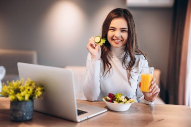 Neşeli genç bir kadın portakal suyu içip kaseden salata yerken bir yandan da mutfakta dizüstü bilgisayardan film izliyor..