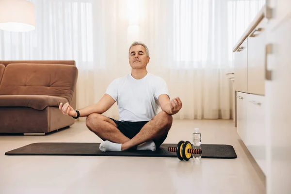 Спортивний зрілий чоловік медитує після практики йоги. Чоловік сидить на чорному йога килимі з закритими очима в сучасній вітальні і розслабляється в легкій позі сидіння. Подбайте про свою концепцію психічного здоров'я . — стокове фото