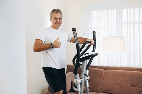 Olgun, mutlu bir adam, eliptik antrenörle pratik yapıyor ve başparmak kaldırıyor. Evde idman yapıyorum. Çapraz antrenör kullanarak kardiyo eğitimi. Arka planda beyaz oturma odası.