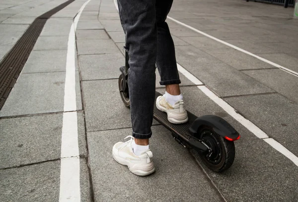 Piernas de tipo anónimo montar scooter eléctrico moderno en camino de asfalto en el fondo borroso de la calle de la ciudad. Concepto de viaje rápido. Elegante joven alquiló un e-scooter. Zapatillas blancas. — Foto de Stock