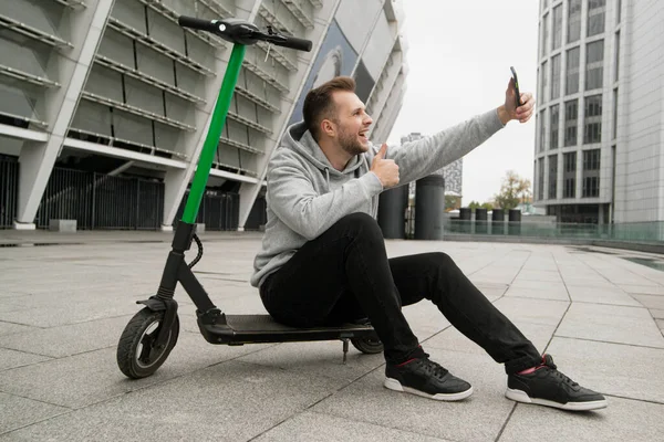 A Guy le gusta el nuevo servicio de alquiler de scooters eléctricos. Hace videollamadas a sus amigos y habla sobre los beneficios de esta aplicación para teléfonos inteligentes. El hombre se sienta en e-scooter, toma selfie y pulgares hacia arriba. — Foto de Stock