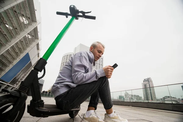 Stijlvolle man zit op een e-scooter en wacht op telefoontje van de verhuurdienst. Milieuvriendelijk vervoersconcept. Wachtconcept. Grijs appartement blokken op de achtergrond. — Stockfoto