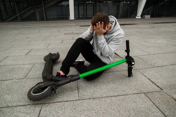 Wees voorzichtig Guy viel tijdens het rijden op de elektrische scooter snel. Man met grijze capuchon zit op de grond en heeft hoofdpijn. Milieuvriendelijk vervoersconcept. Moderne technologieën. Hersenschudding. — Stockfoto